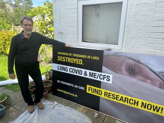 Mark Harper, Chair of our group, standing in a garden next to a very large poster (2m by 1m) due to be displayed on his car. The poster says 'Hundreds of thousands of lives destroyed, Long Covid and ME/CFS' No Treatments, No cure - fund research now!' and links to this website). 