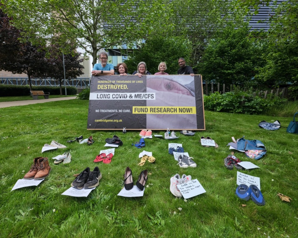 Group of 5 people standing behind the billboard demanding research and treatment for people with ME and Long Covid - in front rows of shoes with names of people absent as too ill to attend.
