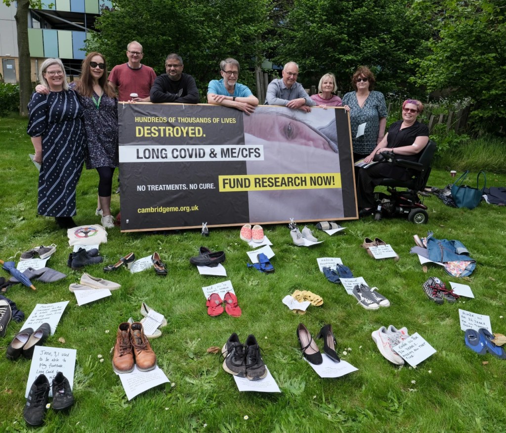Group of 9 people standing behind the billboard demanding research and treatment for people with ME and Long Covid - in front rows of shoes with names of people absent as too ill to attend.