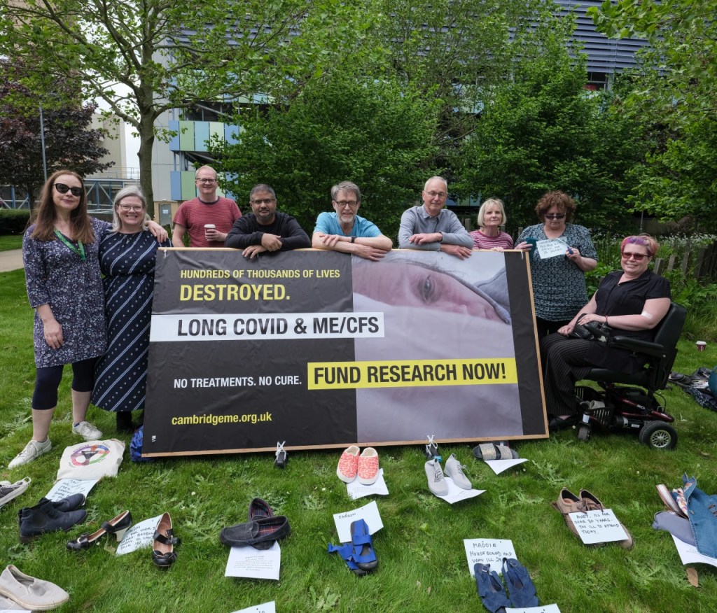 Group of 9 people standing behind the billboard demanding research and treatment for people with ME and Long Covid - in front rows of shoes with names of people absent as too ill to attend.