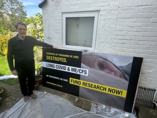Mark Harper, Chair of Cambridge ME Group, in his garden, standing next to large poster propped against a wall.